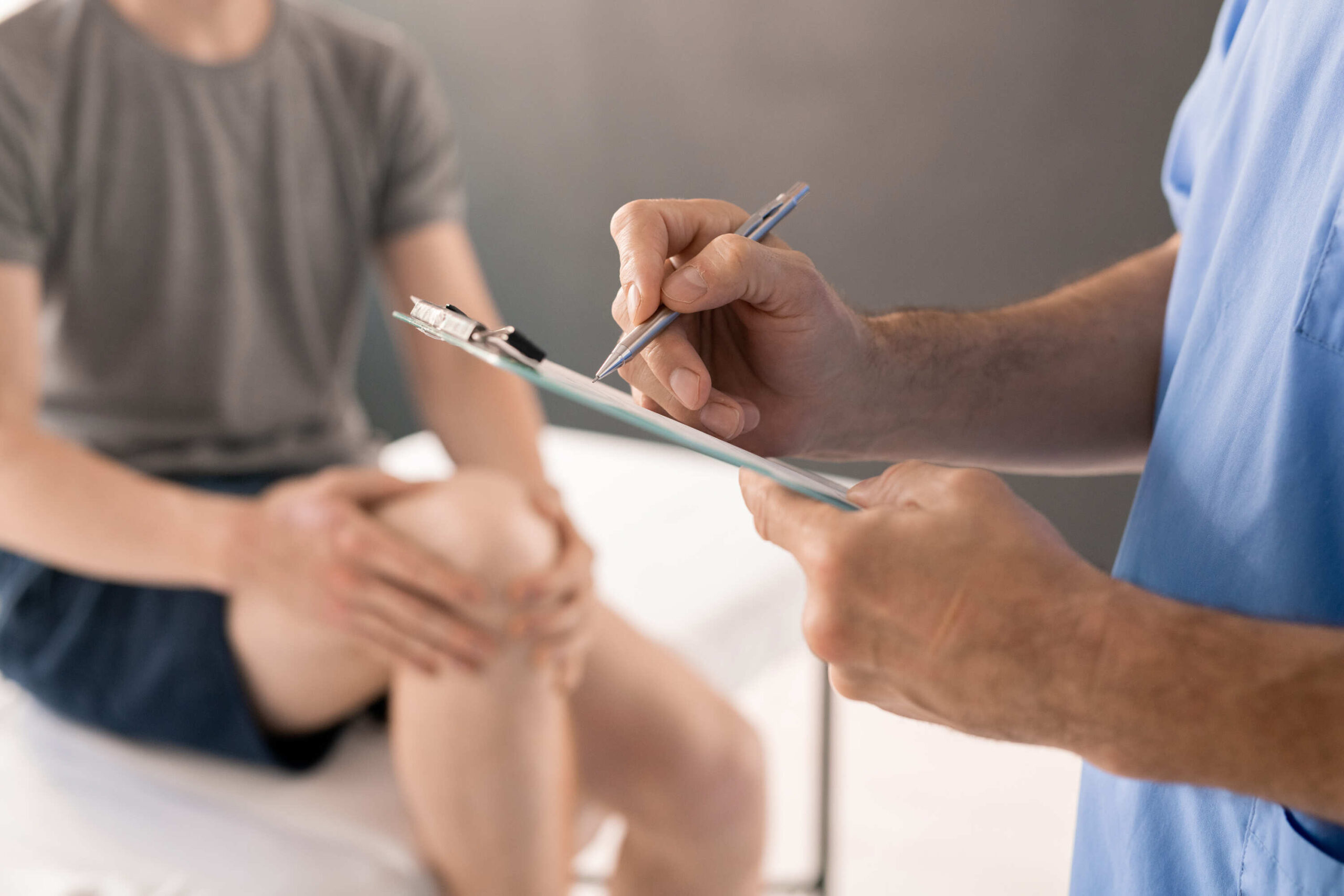 A medical professional takes notes while evaluating a patient who is sitting on a treatment table with their hand on their knee.