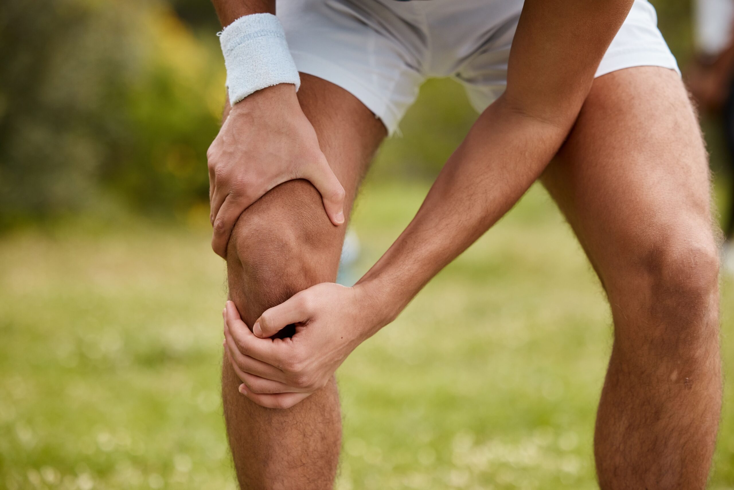 A person in athletic shorts is holding their knee, indicating discomfort while standing on grass.