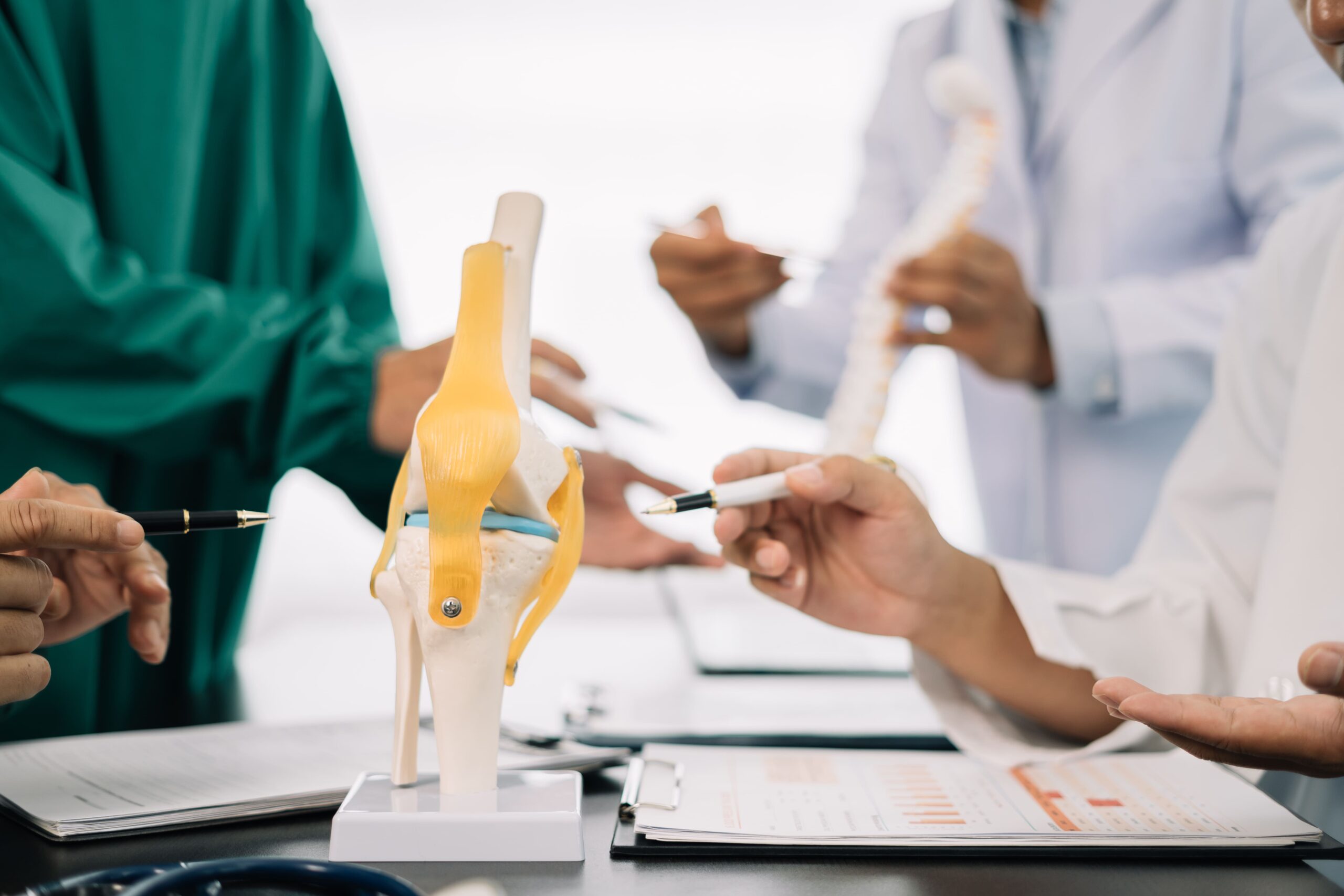 A group of medical professionals discusses a knee joint model while examining documents on a table.