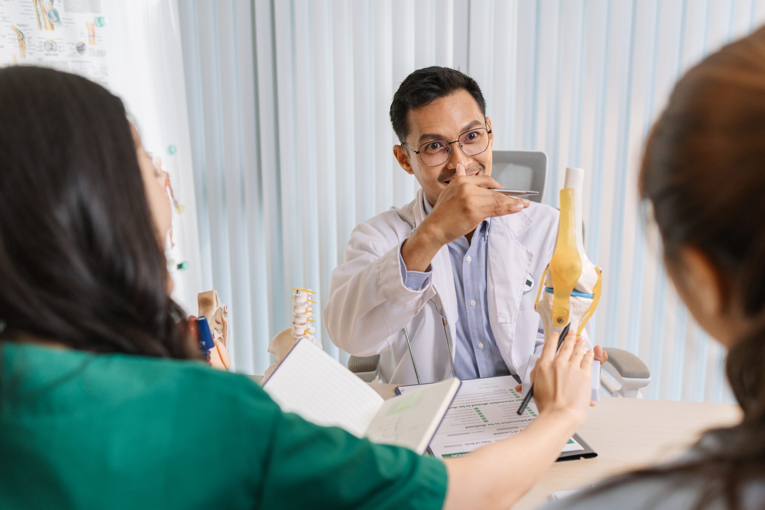 A doctor explains a medical concept using a knee joint model to two attentive patients during a consultation.