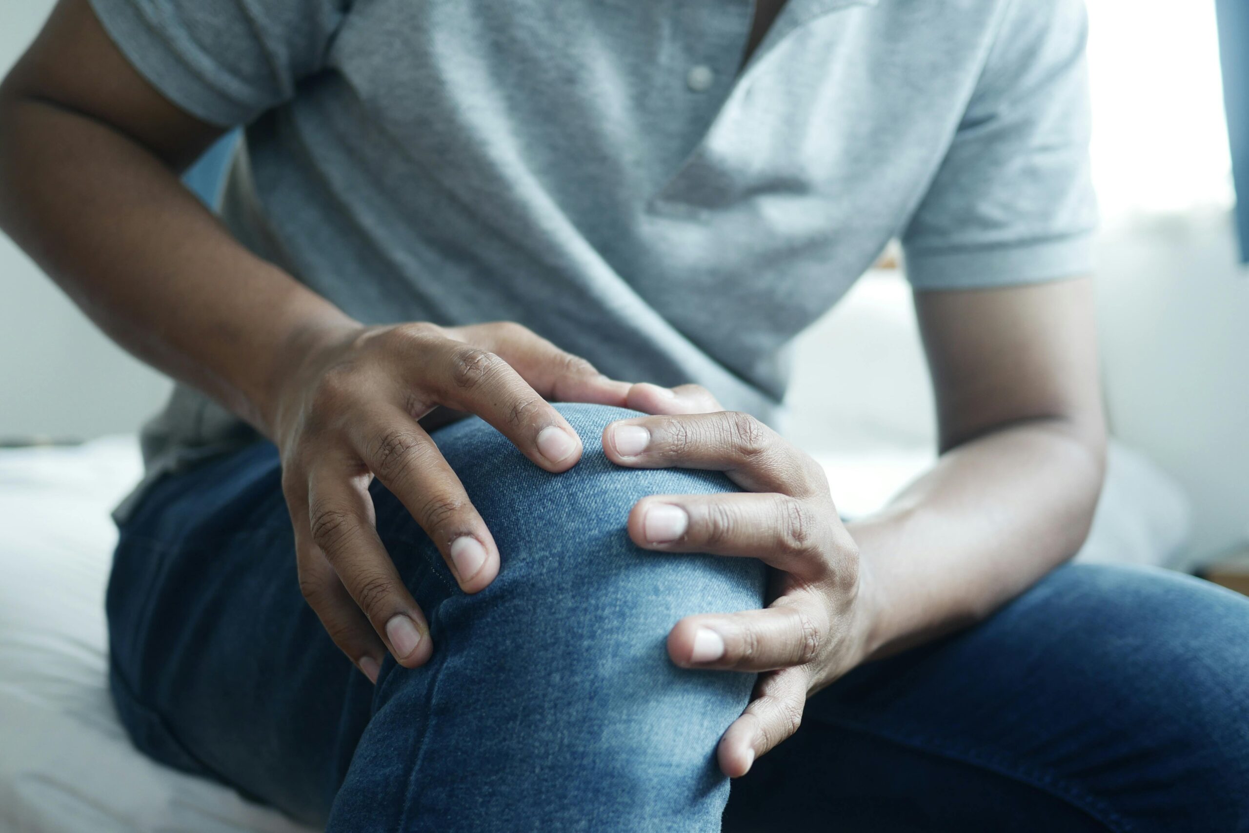 A person with dark skin is seated and holding their knee in discomfort while wearing a gray polo shirt and blue jeans.