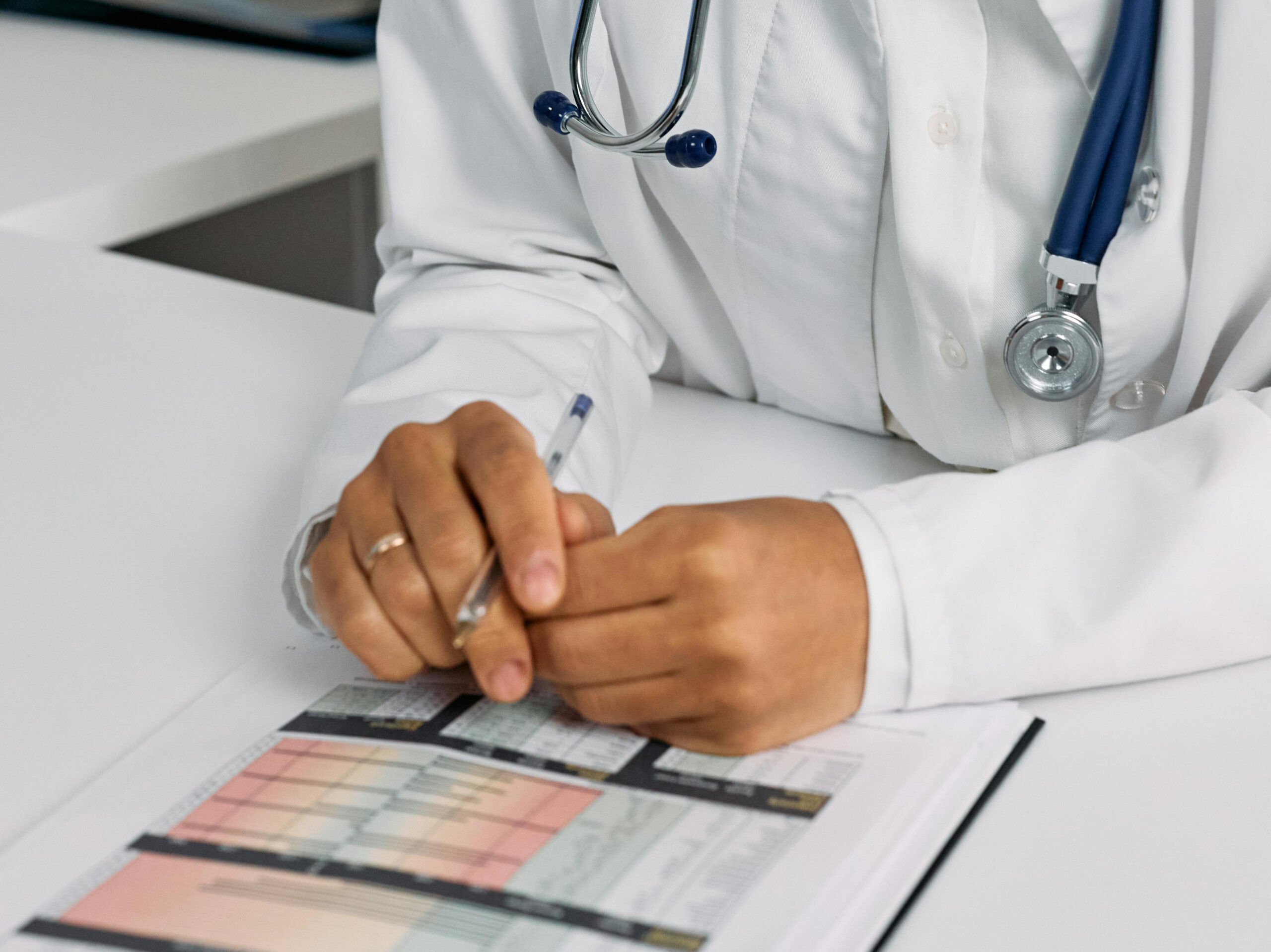 A close-up of a person's hands, wearing a white coat with a stethoscope around their neck, holding a pen over a document with charts.