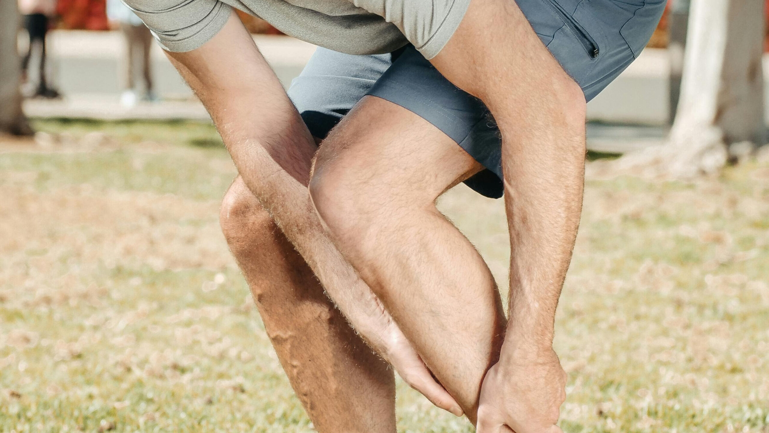 A person in athletic clothing is bending down and holding their ankle in a grassy area.