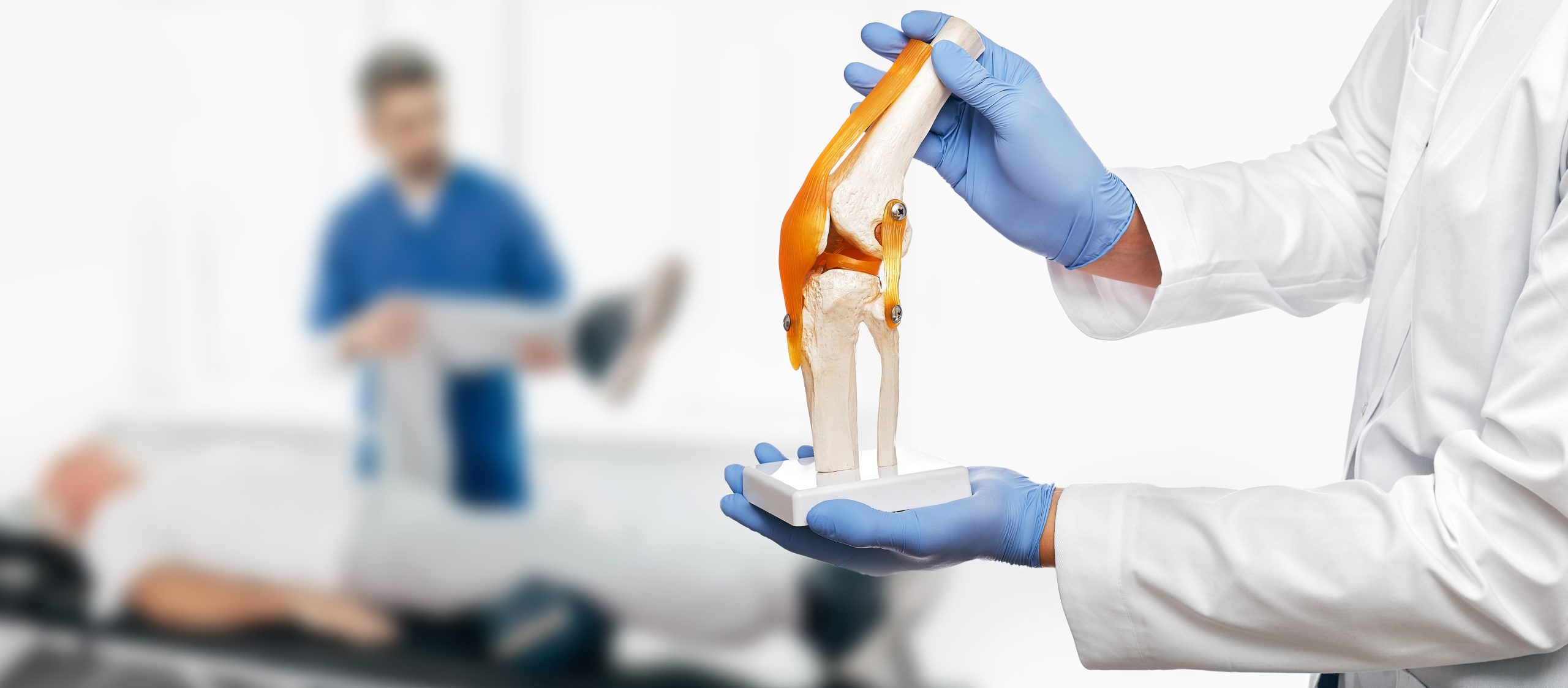 A medical professional in a white coat holding an anatomical model of a knee joint, with a patient lying on a treatment table in the background.