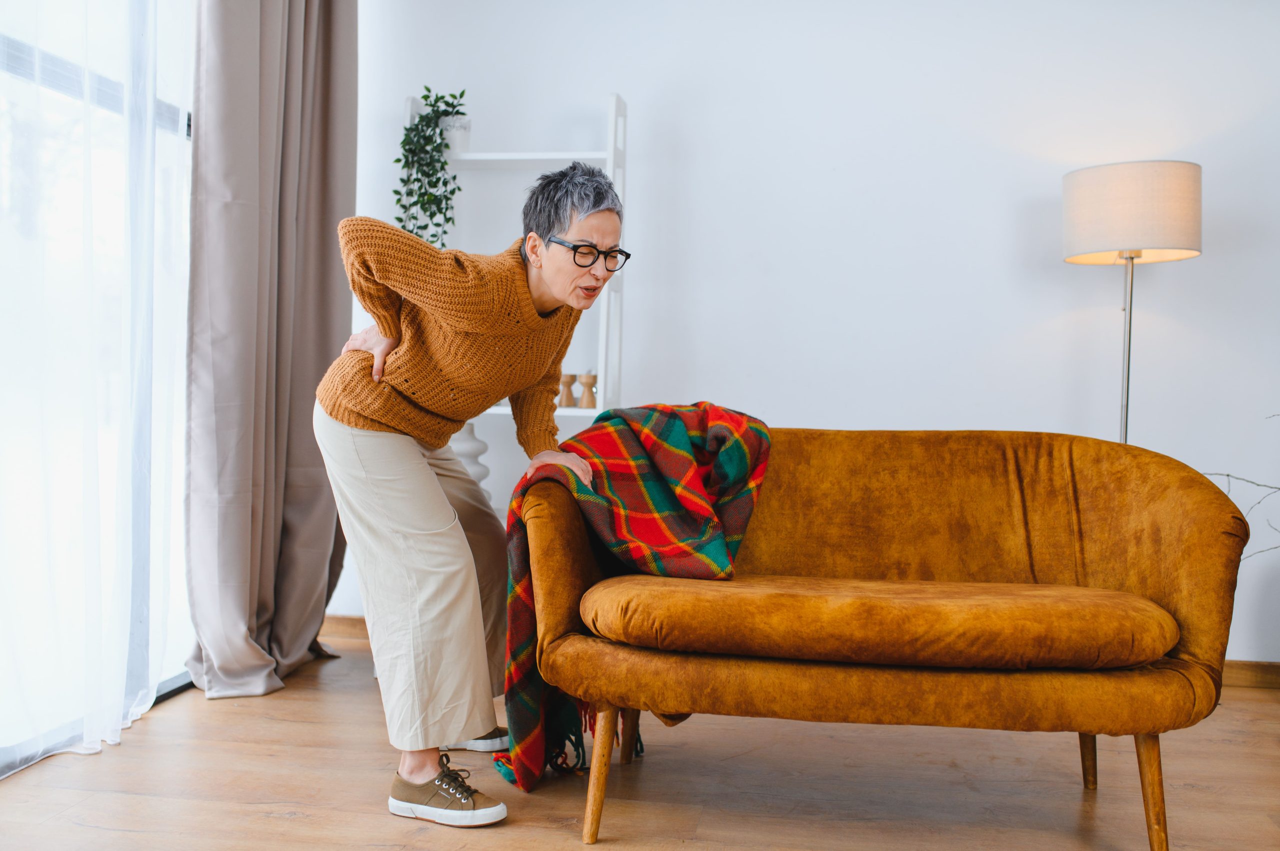 A woman experiencing back pain while leaning on a sofa in her living room.